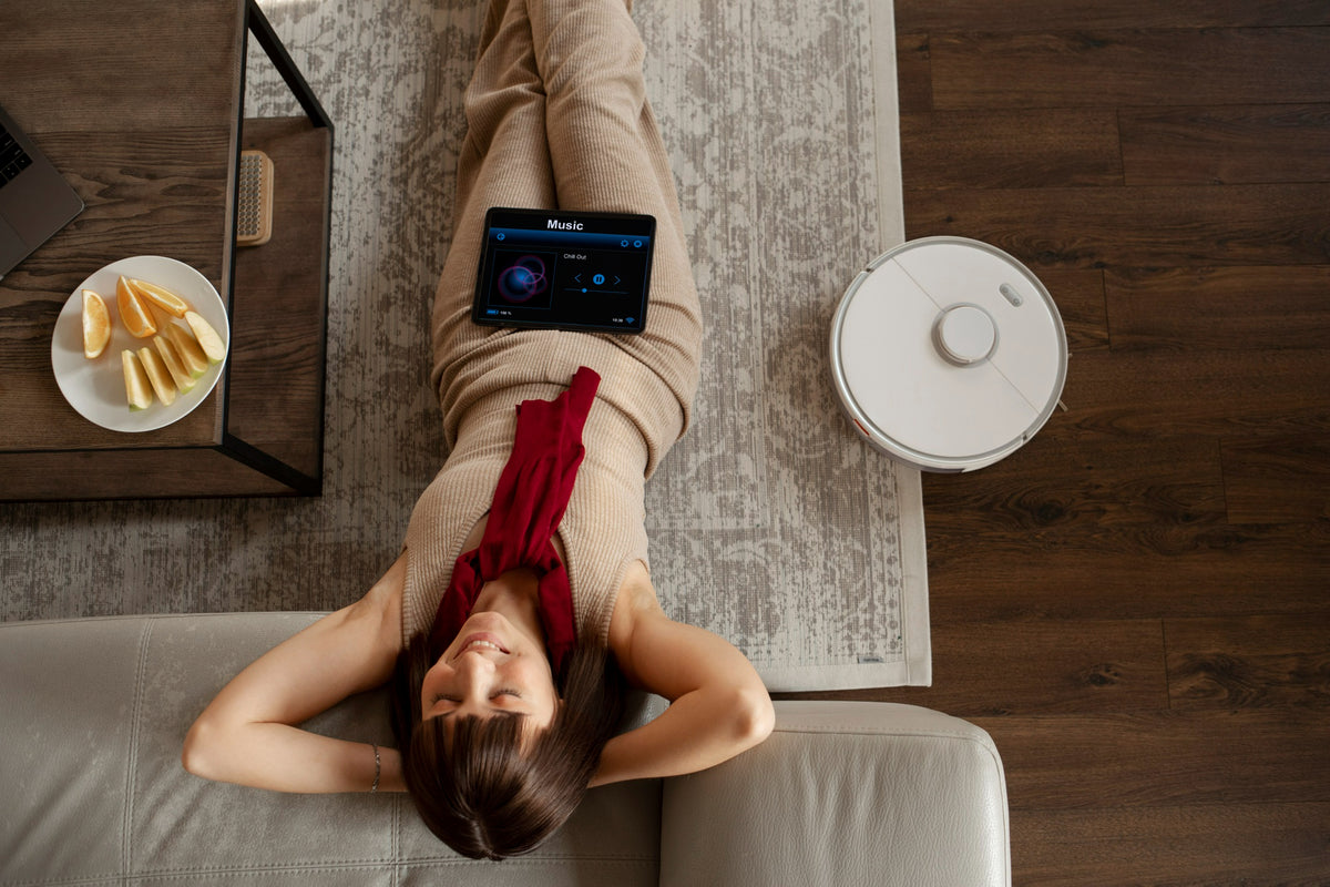 Person lying on a couch with a tablet and a robot vacuum cleaner in a room.